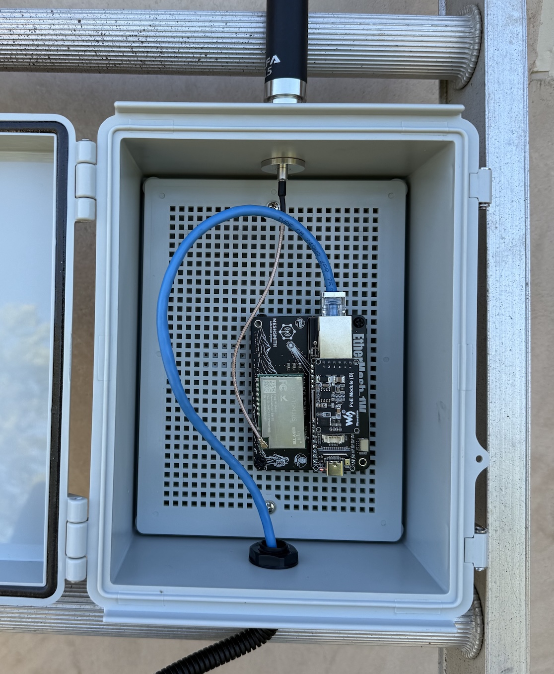 Plastic enclosure containing a MeshSmith EtherMesh-1W, connected via an ipex to a N-Type bulkhead connector with an Alfa Antenna exiting the top. Blue Ethernet enters through a waterproof gland at the bottom, covered in Nylon conduit on the exterior.